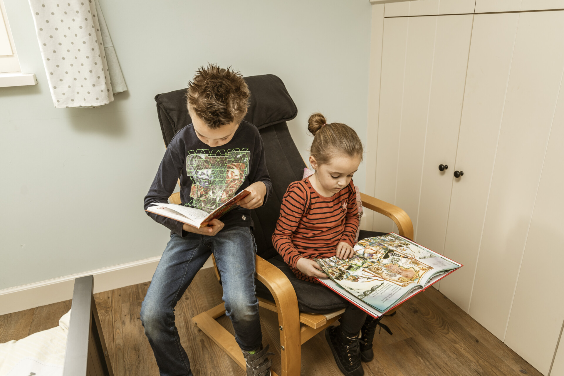 Twee jonge kinderen zitten samen in een stoel en lezen aandachtig ieder hun eigen boek uit het Boekskes-pakket in een knusse, rustige kinderkamer.