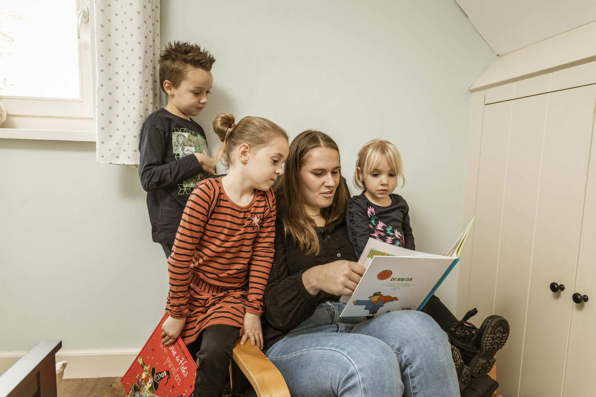 Moeder leest voor aan drie jonge kinderen op een knusse kamer; iedereen kijkt aandachtig naar het prentenboek Dikkie Dik.
