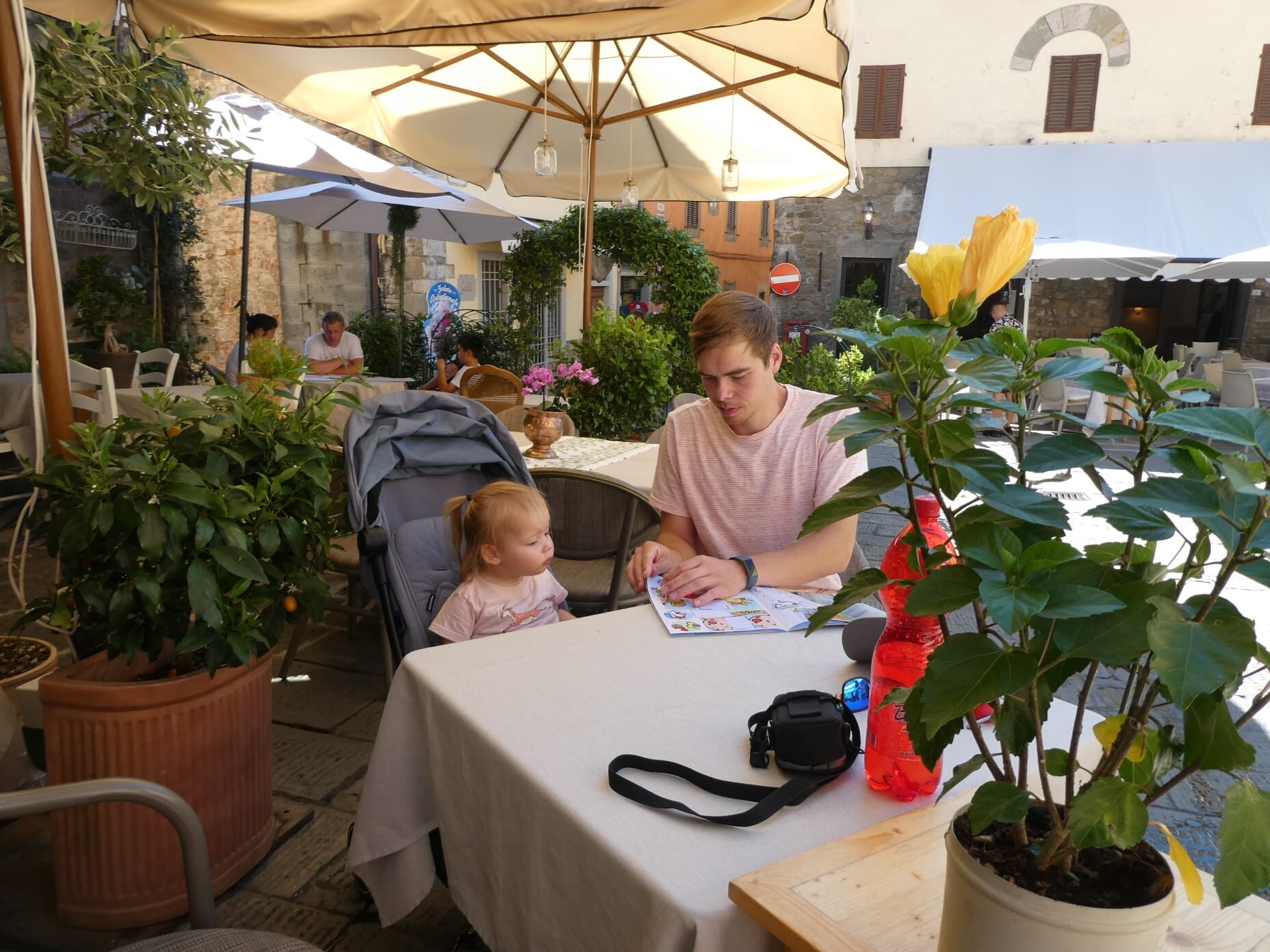 Vader leest een kinderboek uit het Boekskes Peuterpakket voor aan zijn dochter op een zonnig terras in een knusse, groene straat in een historisch stadje.