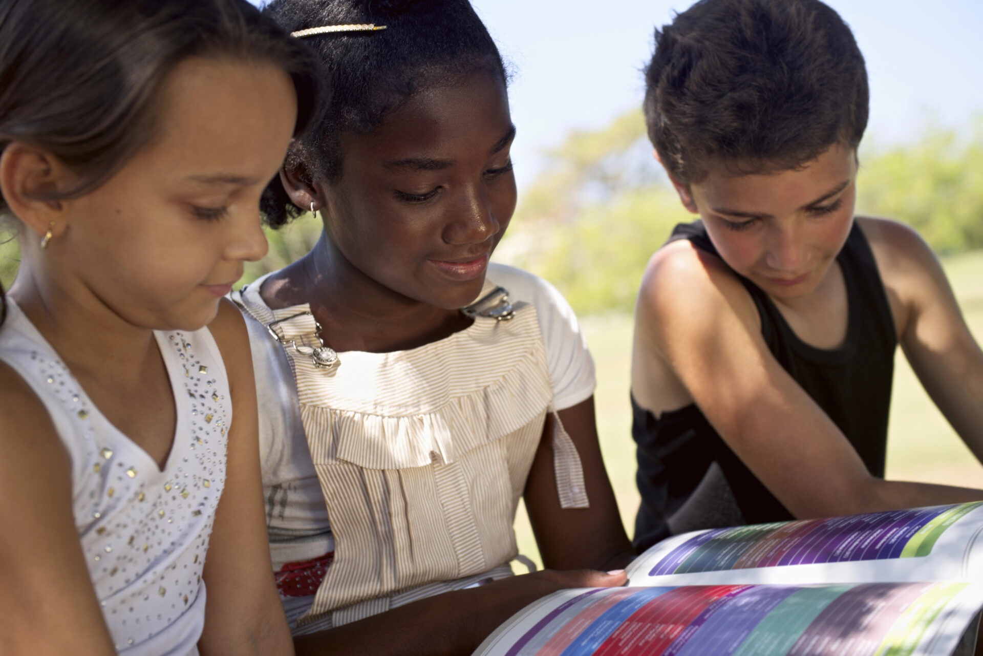 Drie kinderen lezen samen buiten in een kleurrijk kinderboek en genieten van een zonnige vakantiedag.