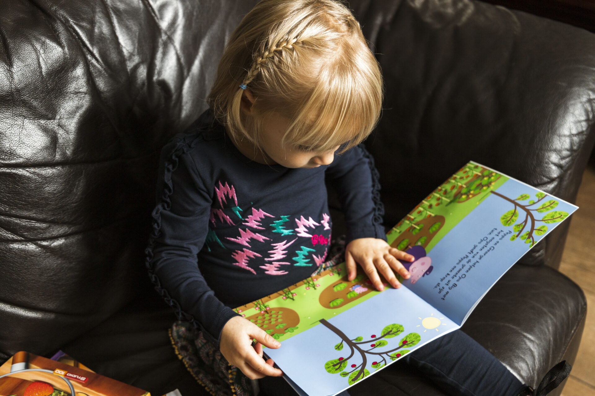 Meisje zit op de bank en bladert aandachtig door een kleurrijk prentenboek vol vrolijke illustraties uit het Boekskes-pakket van december.